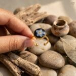 A dainty golden ring featuring a blue flower set against a dark resin background